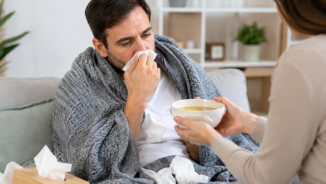 Sick man wrapped in blanket holding tissue while receiving bowl of soup for flu relief at home. concept of comfort during illness, caring support, home remedy for cold
