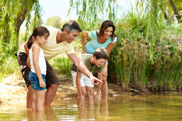 Obraz premium Happy, parents and pointing with children at lake, adventure and discover fish on holiday. Smile, people and hand gesture in nature with kids, river observation and water for sightseeing vacation