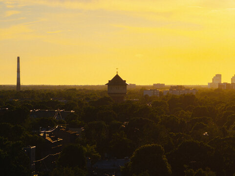 Aerial view of the historic water tower surrounded by lush green trees and urban buildings under a warm golden hour sky in Riga, Latvia.