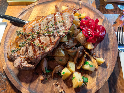 High angle close-up showing grilled meat steak topped with thyme, served with roasted potato, mushroom, and pickled red onion on round wooden board