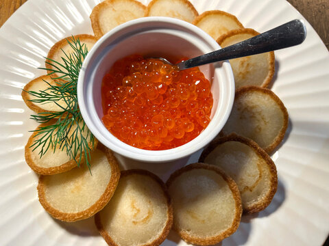 Bright orange salmon roe caviar served in a white bowl with a spoon, surrounded by golden blinis arranged in a circle and fresh dill on a white plate