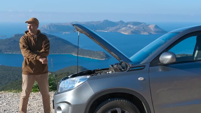 Stressed driver checking under the hood of a broken car. Frustrated man standing on a scenic coastal road trying to figure out what is wrong with his broken-down car, inspecting the engine