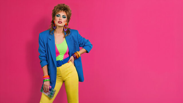 A woman in vibrant 1980s fashion, featuring a blue blazer, yellow pants, and bold makeup, poses against a solid pink background with ample copy space on the right.