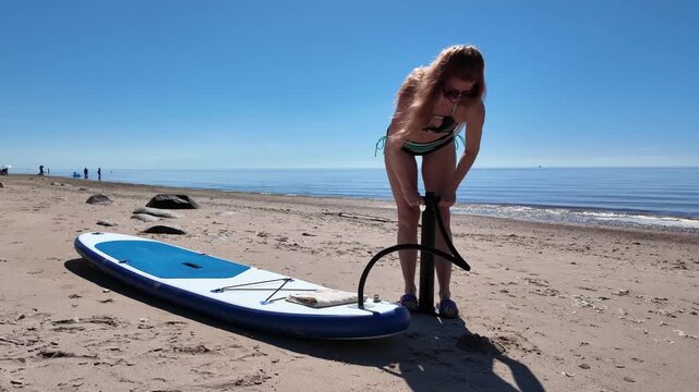 Woman inflating stand up paddle board with hand pump on sunny beach before paddling