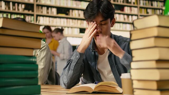 Tired asian student leaning over open book between tall stacks in library, studying alone near multiple textbooks at wooden table study area, Literature self-study indoors, supporting study session.