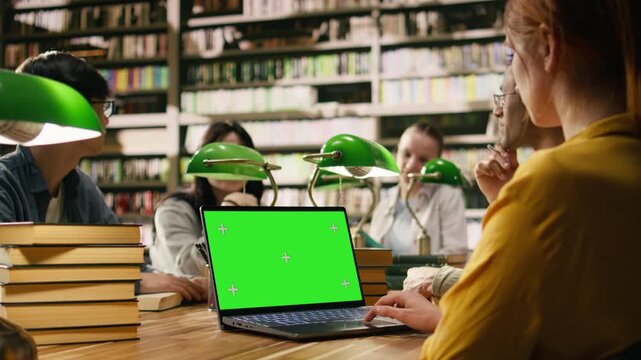 Green screen mockup on laptop with peers at wooden library desk, student hand uses the touchpad near editable screen, Modern higher education with chroma key blank screen, supporting group study.