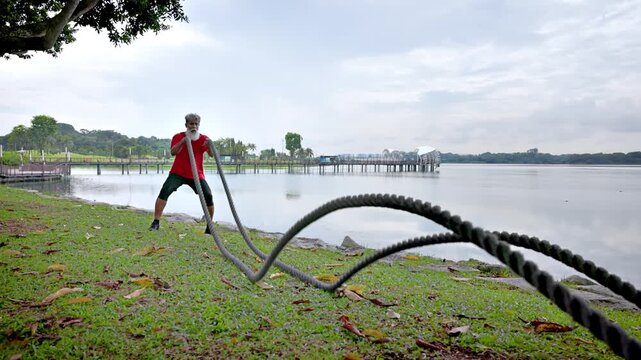 Mature Indian man with a beard working out with a battle rope at a lakeside park In Singapore. Slow motion.