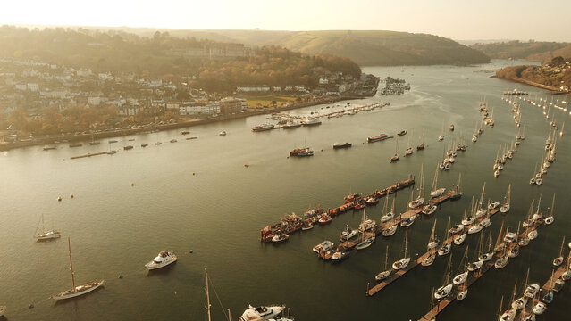 Aerial view of Kingswear marina with sailboats and yachts moored at wooden pontoons, surrounded by rolling green hills and coastal buildings Kingswear, England, United Kingdom.