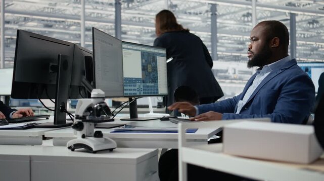 Man examining microchip circuit layouts on monitor in factory R&D office using specialized software tools, African American worker improving semiconductor manufacturing used in smart devices.