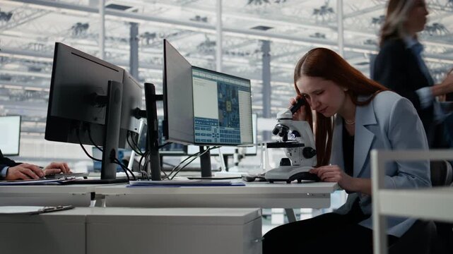 Woman in microelectronics development facility studying microchip architecture with microscope, female engineer using microscopy tool to research semiconductor materials in office.
