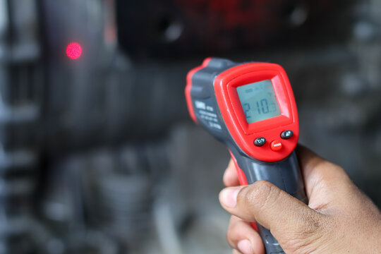 Close-up of a hand holding a digital infrared laser thermometer measuring engine temperature in a workshop.
