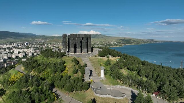 Drone shot capturing the massive bronze and stone pillars of the Chronicle of Georgia monument complex, situated on a green hill overlooking the vast Tbilisi Sea reservoir under a clear blue sky