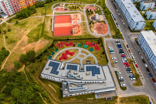 Aerial view of a modern building with solar panels on the roof, adjacent to a colorful playground, a sports court, and a parking lot in a residential area of Brno, South Moravian Region, Czechia.