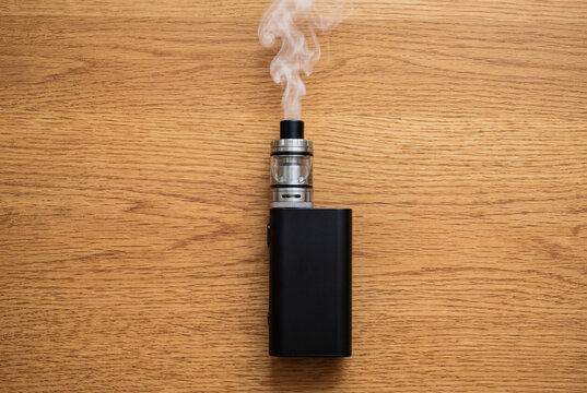 Black vape mod electronic cigarette device producing vapor on a wooden table