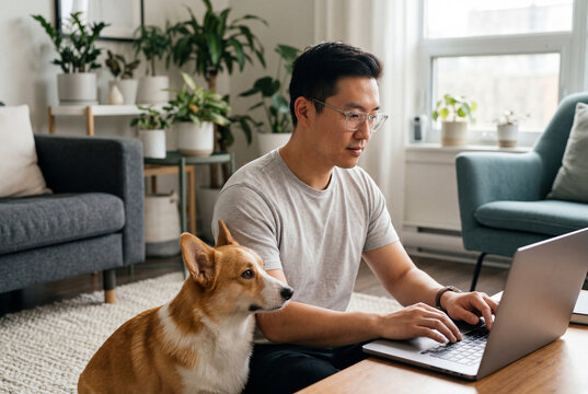 Asian man using a laptop while sitting on the floor next to his corgi dog
