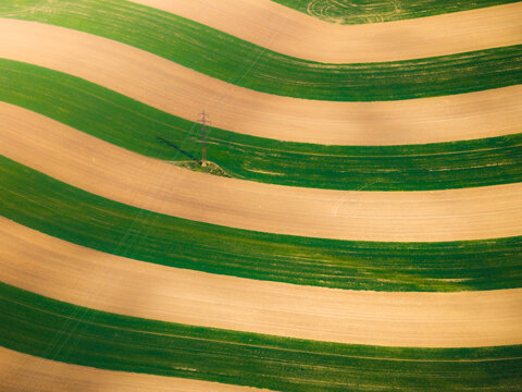 Aerial view of striped agricultural fields with alternating green and brown soil patterns and a single power line tower in Brno, South Moravian Region, Czechia.