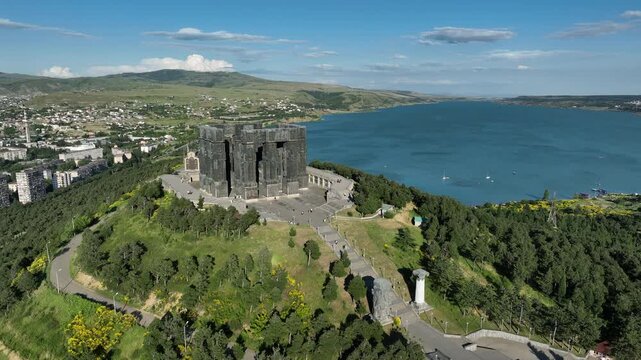 Drone shot capturing the massive bronze and stone pillars of the Chronicle of Georgia monument complex, situated on a green hill overlooking the vast Tbilisi Sea reservoir under a clear blue sky