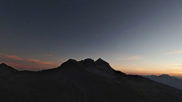 Aerial view of the French Alps mountain range silhouetted against a twilight sky with a crescent moon visible above the peaks in Chamonix, Auvergne-Rhone-Alpes, France.