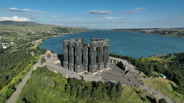 Drone shot capturing the massive bronze and stone pillars of the Chronicle of Georgia monument complex, situated on a green hill overlooking the vast Tbilisi Sea reservoir under a clear blue sky