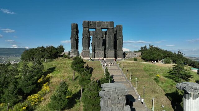 Drone shot capturing the massive bronze and stone pillars of the Chronicle of Georgia monument complex, situated on a green hill overlooking the vast Tbilisi Sea reservoir under a clear blue sky
