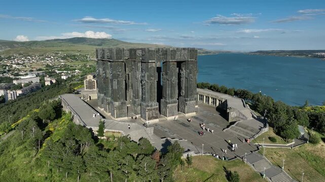Drone shot capturing the massive bronze and stone pillars of the Chronicle of Georgia monument complex, situated on a green hill overlooking the vast Tbilisi Sea reservoir under a clear blue sky