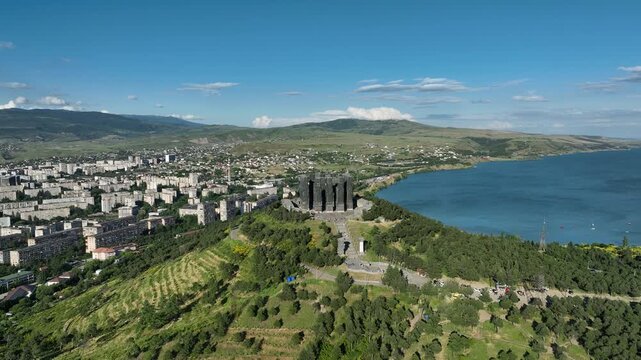 Drone shot capturing the massive bronze and stone pillars of the Chronicle of Georgia monument complex, situated on a green hill overlooking the vast Tbilisi Sea reservoir under a clear blue sky
