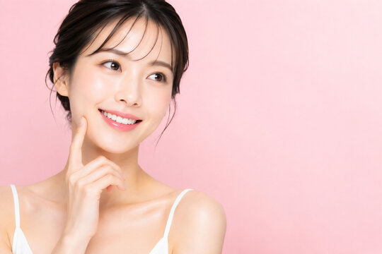ピンクの無地背景で白い歯を見せて微笑みながら頬に指を添える若い女性の明るく清潔感ある美容イメージ