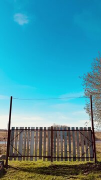 Weathered metal gate with vertical stainless strips, chain link fences flanking sides, spring grass carpeting ground, clear blue