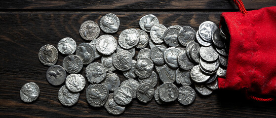 Ancient silver coins spilling out of a red pouch onto a dark rustic wooden table. Roman denarius treasure hoard flat lay. Panoramic banner © scaliger