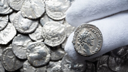 Gloved hand holding an ancient Roman silver coin of Emperor Commodus. Macro close up of a numismatist examining a historical artifact. Pile of old collection in blurred background © scaliger