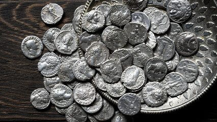 Top view of ancient Roman and Greek silver coins spilling from an ornate metal dish onto a dark wooden table. Historical money collection and antique treasure concept © scaliger