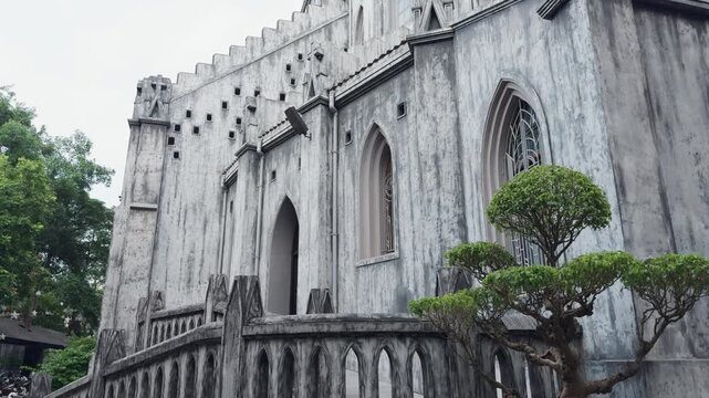 Grey stone church building with its weathered facade and narrow arched windows, featuring gothic revival architectural elements and a small, vibrant green tree in the foreground