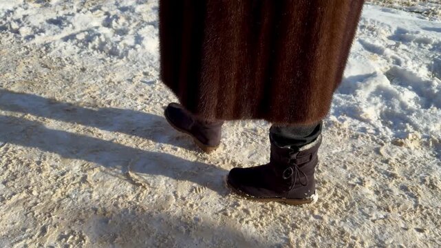 Close-up woman's suede boots walking through snow, mink coat hem visible, frost sparkling on powdery ground, steady steps leaving