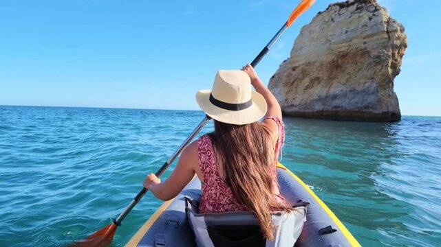 Rear view of a woman with long hair and a sun hat kayaking on a beautiful sunny day, exploring the stunning coastline with picturesque rock formations and clear blue water of the atlantic ocean