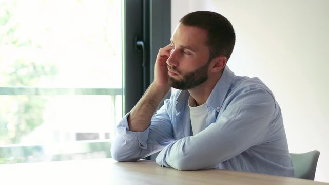 Young handsome man with a beard sitting alone at a table, looking out the window while lost in thought and feeling pensive, reflecting on life's problems and feeling lonely