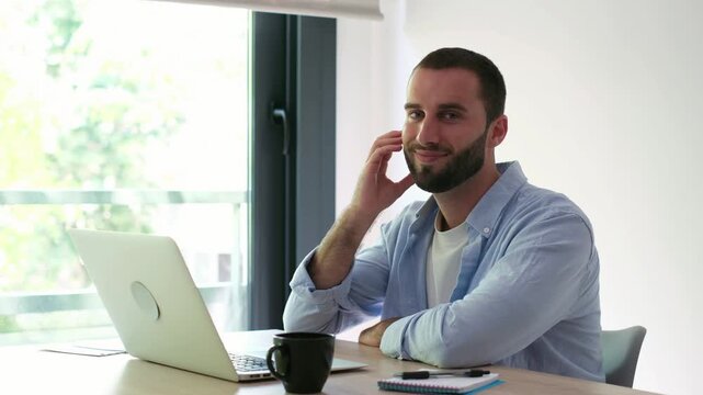 Successful young businessman with a beard sitting at his desk with a laptop and smiling at the camera, showcasing confidence and satisfaction with his work from a bright home office