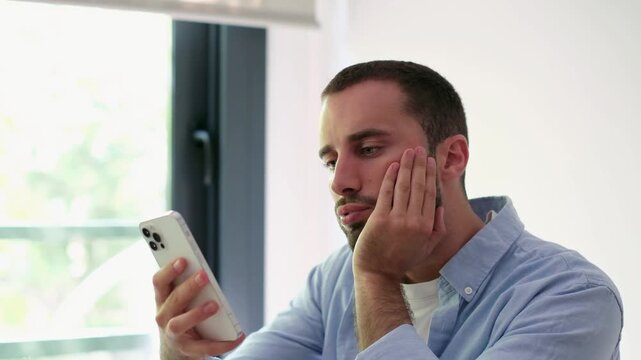 Young handsome man feeling tired and bored while endlessly scrolling through social media on his smartphone, showing clear signs of digital fatigue and procrastination at home
