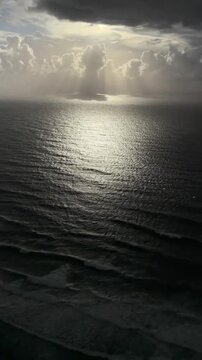 Radiant sunbeams penetrate through heavy dark storm clouds above the vast deep blue ocean surface creating a dramatic shimmering light reflection on rippling water waves in this cinematic high definit