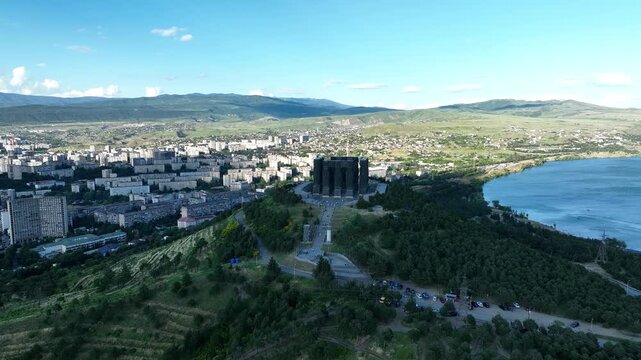 Drone shot capturing the massive bronze and stone pillars of the Chronicle of Georgia monument complex, situated on a green hill overlooking the vast Tbilisi Sea reservoir under a clear blue sky