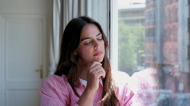 Thoughtful young woman with a pensive expression standing by the window at home, gazing outside while contemplating life. A moment of introspection and reflection on a sunny day