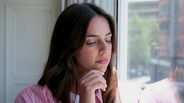 Thoughtful and contemplative young woman with her hand on her chin looking out the window, lost in thought and reflecting about life. Beautiful brunette girl pondering and daydreaming at home