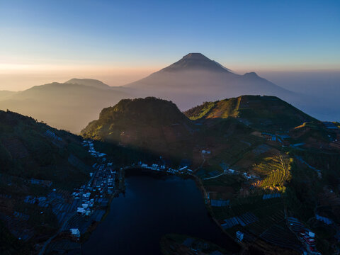 Aerial view of Telaga Cebong lake surrounded by terraced agricultural fields and hills at sunrise with Mount Sikunir in the background in Kejajar, Central Java, Indonesia.
