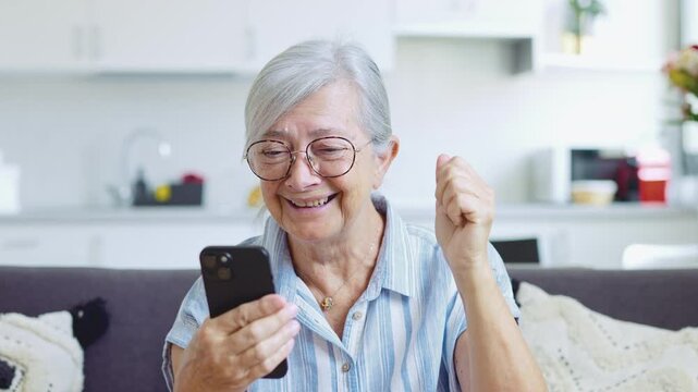 Cheerful senior woman sitting on the sofa at home, using her smartphone and celebrating success with a fist pump after receiving exciting news, winning an online game, or getting a great offer