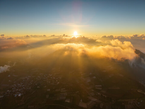 Aerial view of the Dieng plateau at sunrise with golden light illuminating the clouds above terraced agricultural fields in Kejajar, Central Java, Indonesia.