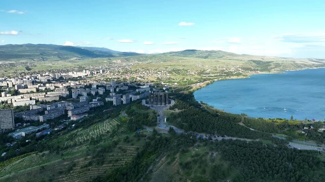 Drone shot capturing the massive bronze and stone pillars of the Chronicle of Georgia monument complex, situated on a green hill overlooking the vast Tbilisi Sea reservoir under a clear blue sky