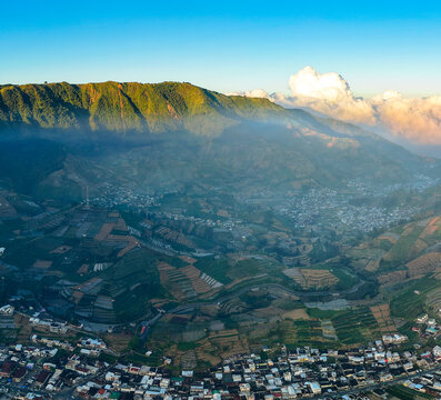 Aerial view of the Dieng Plateau featuring terraced agricultural fields and village houses nestled among misty mountains in Kejajar, Central Java, Indonesia.