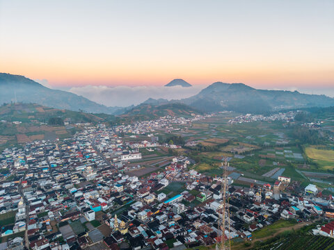 Aerial view of Dieng plateau town nestled among lush green agricultural fields and rolling mountains under a soft sunrise sky in Kejajar, Central Java, Indonesia.