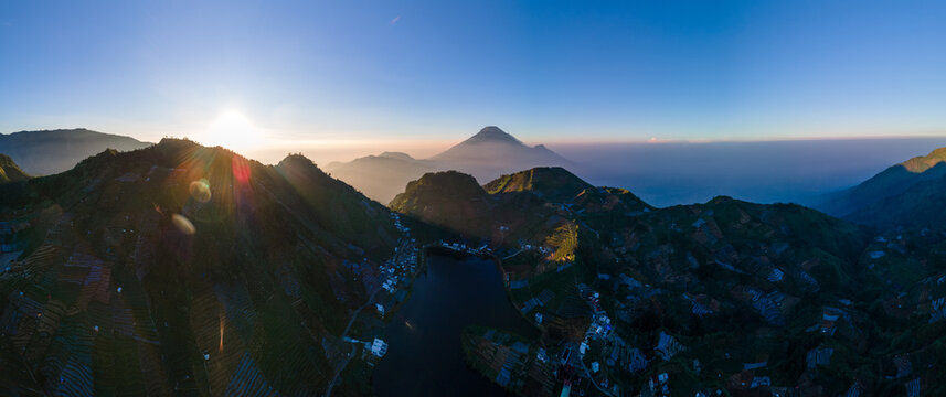 Aerial view of Telaga Cebong lake surrounded by terraced agricultural fields and mountain slopes at sunrise in Kejajar, Central Java, Indonesia.