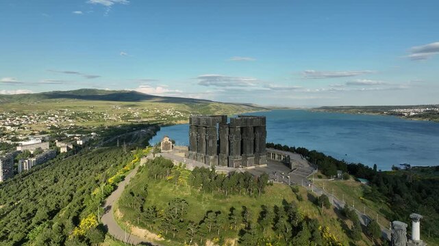 Drone shot capturing the massive bronze and stone pillars of the Chronicle of Georgia monument complex, situated on a green hill overlooking the vast Tbilisi Sea reservoir under a clear blue sky