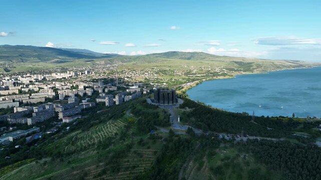Drone shot capturing the massive bronze and stone pillars of the Chronicle of Georgia monument complex, situated on a green hill overlooking the vast Tbilisi Sea reservoir under a clear blue sky